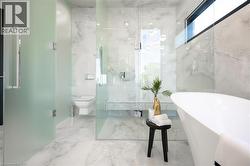 Bathroom featuring a freestanding white soaking tub, large format marble-look wall and floor tiling, a walk-in shower with a built-in bench, and a privacy glass enclosure for the commode - 