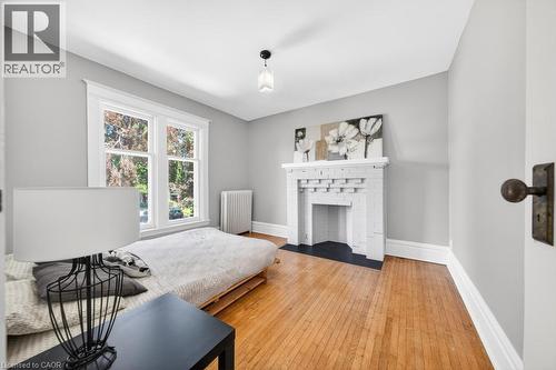 Bright room featuring a decorative fireplace with a black hearth, hardwood flooring, and a large double window with white trim - 35 Burris Street, Hamilton, ON - Indoor Photo Showing Bedroom With Fireplace