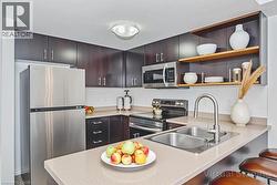 Modern kitchen featuring stainless steel appliances, dark wood cabinetry, light-toned countertops, a dual basin sink with a pull-down faucet, and integrated open shelving - 