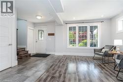 Open-concept living area featuring wood-finish flooring, a decorative glass front door, and a multi-pane window with exterior greenery - 