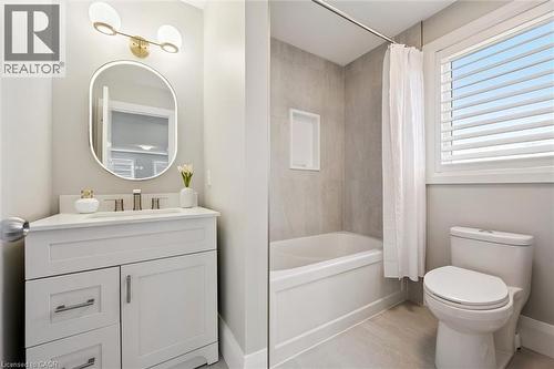 Bathroom featuring a white vanity with built-in sink, an illuminated oval mirror, a full bathtub with a tiled surround and recessed shelf, and a toilet - 72 Elmbank Trail, Kitchener, ON 
