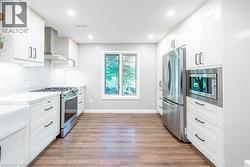 Kitchen with appliances with stainless steel finishes, light wood-type flooring, light stone counters, wall chimney range hood, and backsplash - 