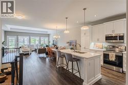 Open-concept living space featuring wood-finish flooring, a contemporary kitchen island with seating, white shaker cabinetry, stainless steel appliances, and abundant natural light from multiple windo - 