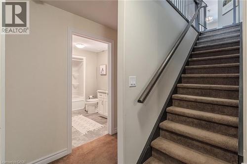 Carpeted staircase with dark handrail and balusters, featuring a doorway leading to a bathroom with a white vanity, toilet, and tub/shower combination - 15 Winterberry Lane, Waterford, ON 