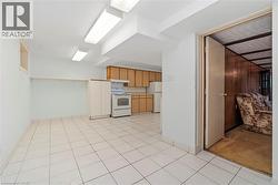 Spacious area featuring ceramic tile flooring, a kitchen zone with wood-finish cabinetry and white appliances, fluorescent overhead lighting, and an open doorway revealing a separate space with wood p - 