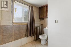 Bathroom featuring tiled walls, a window with curtain rod, a toilet, a radiator, and a wall-mounted wooden cabinet - 