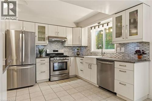 Kitchen featuring stainless steel appliances, white cabinetry, a farmhouse sink, granite-finish countertops, and a decorative tile backsplash - 17 Piper Place, Caledonia, ON 