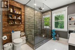 Glass-enclosed shower with stacked stone-finish tile surround, exposed brick accent wall, white-framed window, built-in shelving, and white floor tiling - 