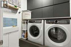 Dedicated laundry area featuring front-loading washer and dryer units, a wall-mounted utility sink with blue subway tile backsplash, dark flat-panel cabinetry, and a light wood-finish shelf - 