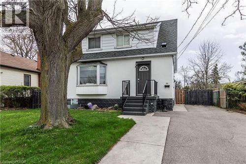 White stucco exterior with a dark shingled roof and a prominent mature tree in the front yard - 20 West 1St Street, Hamilton, ON 