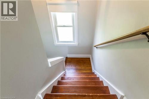 Staircase featuring wood-finish treads, a wood handrail with dark metal brackets, light neutral wall paint, white trim, and a double-hung window with white framing - 20 West 1St Street, Hamilton, ON 