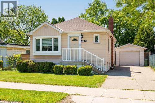 Residential exterior featuring a light-colored siding, a front entry porch with white railings, a prominent bay window, a brick chimney, and a detached garage with a driveway - 420 Upper Gage Avenue, Hamilton, ON 