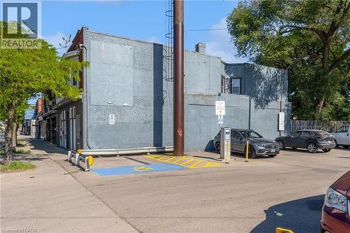 Street-level building featuring a brick exterior with boarded-up windows, a corner storefront with multiple window displays, and a paved parking area with designated accessibility spaces - 1371 Main Street E, Hamilton, ON - Outdoor