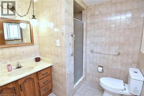 Bathroom featuring a walk-in shower with a frosted glass door, ceramic tile walls with decorative accents, an oak-finish vanity with an integrated sink, a stone-finish countertop, and a wall-mounted m - 372 Fennell Avenue W, Hamilton, ON 