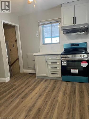 Kitchen area featuring wood-finish flooring, light-colored cabinetry with dark hardware, a freestanding range with an overhead vent, and a window with blinds - 102 Massey Avenue, Kitchener, ON 