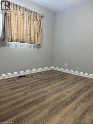 Spacious room featuring wood-finish flooring, light grey painted walls, white baseboards, and a window with curtains - 102 Massey Avenue, Kitchener, ON 