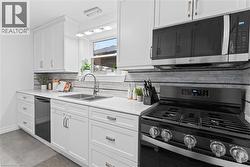 Modern kitchen featuring white shaker cabinetry, light-toned countertops, a stainless steel double basin sink with a gooseneck faucet, a tile backsplash, and a black gas range with a coordinating micr - 