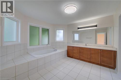 Spacious bathroom featuring a built-in soaking tub surrounded by white tile, multiple frosted windows, and a wood-finish vanity with an integrated sink and an expansive mirror - 180 Arthur Fach Drive, Cambridge, ON 