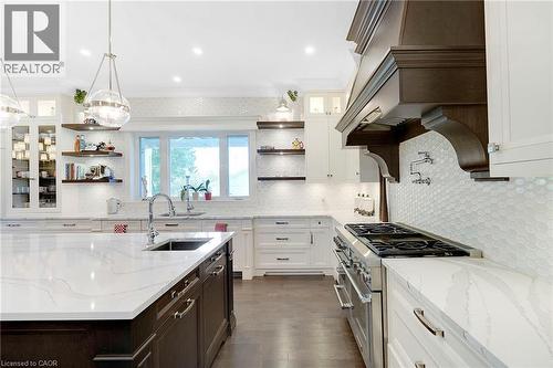 Gourmet kitchen featuring a large island with an undermount sink, white cabinetry, dark wood-finish range hood, white patterned tile backsplash, and stainless steel appliances - 6195 Milburough Line, Burlington, ON - Indoor Photo Showing Kitchen With Upgraded Kitchen