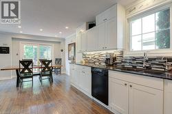 Kitchen and dining area featuring wood-finish flooring, white cabinetry, dark stone countertops, a mosaic tile backsplash, and a double basin sink with a gooseneck faucet - 