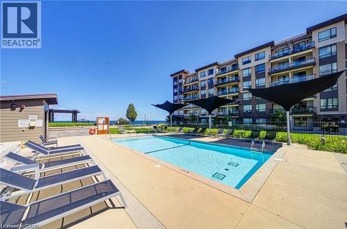 Outdoor swimming pool with concrete surround, sun loungers, and shade structures, set against a multi-story building with balconies and a distant body of water - 40 Esplanade Lane Unit# 614, Grimsby, ON 