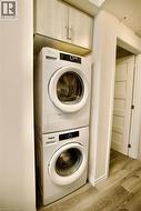 Laundry area featuring a stacked washer and dryer, upper cabinetry with light wood-finish doors, and wood-finish flooring - 