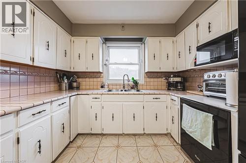 U-shaped kitchen featuring white cabinetry with dark hardware, a tile backsplash, and a double basin sink beneath a window - 70 Webster Road, Hamilton, ON 