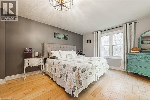 Bright room featuring warm wood-finish flooring, a contemporary ceiling fixture, a tufted headboard, and a large window with white trim - 70 Webster Road, Hamilton, ON 