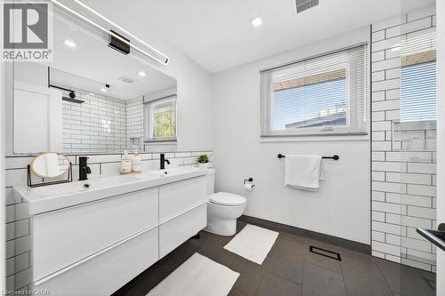 Contemporary bathroom featuring a dual vanity with integrated sinks, matte black fixtures, a large mirror with integrated lighting, a window with horizontal blinds, and subway tile accents - 19 Glen Forest Drive, Hamilton, ON 