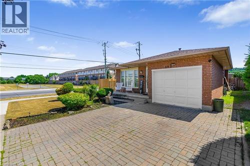 Brick-clad single-story residence featuring an attached garage with a white panel door, a paved driveway, and a concrete front porch - 102 Juniper Drive, Hamilton, ON 