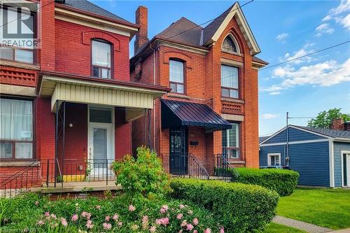 Brick exterior featuring architectural detailing, a covered front porch with wrought iron railing, and a gabled roofline - 244 Hughson Street N, Hamilton, ON 