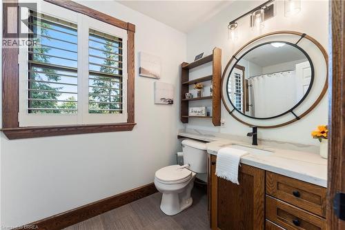 Bathroom featuring a wood-finish vanity with a light-colored countertop, a circular framed mirror, and a window with plantation shutters - 182 Governors Road E, Paris, ON 