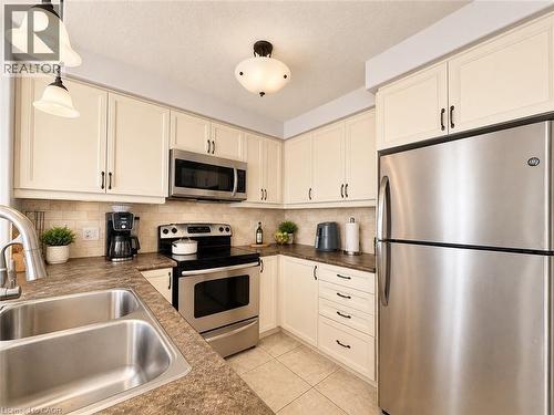 Kitchen featuring off-white cabinetry with dark hardware, a stainless steel double-basin sink, and a tiled backsplash. Staged with AI - 1333 Countrystone Drive, Kitchener, ON 