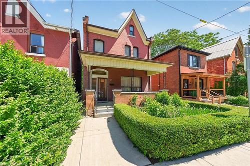 Brick exterior featuring a gabled roofline, covered front porch, and a manicured hedge-lined front garden - 16 Mulberry Street, Hamilton, ON 