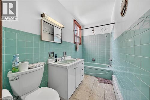 Bathroom featuring teal wall tiling, a white vanity with integrated sink, a white toilet, a built-in bathtub, and a window with wood trim - 16 Mulberry Street, Hamilton, ON 