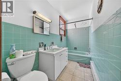 Bathroom featuring teal wall tiling, a white vanity with integrated sink, a white toilet, a built-in bathtub, and a window with wood trim - 