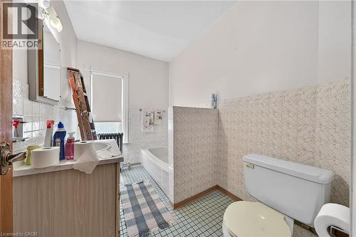 Bathroom featuring a vanity with an integrated sink, a built-in bathtub, a toilet, tiled walls, and a window with a roller shade - 16 Mulberry Street, Hamilton, ON 