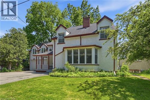 Stucco exterior residence featuring a prominent bay window, multiple gabled dormers, a covered front porch with white railings, a brick chimney, and an attached two-car garage - 3 Ancaster Street E, Dundas, ON - Outdoor With Facade