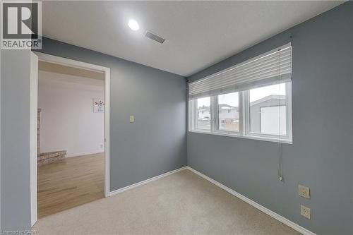 Carpeted room featuring a large window with horizontal blinds, recessed ceiling lighting, and a doorway opening to a space with wood-finish flooring and a partial brick-faced structure - 233 Benesfort Crescent, Kitchener, ON 