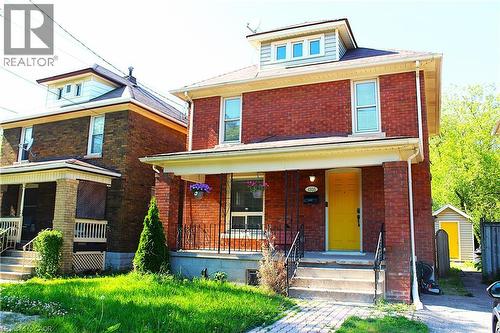 Red brick exterior featuring a bright yellow front door, front porch with columns, dormer window, and a small front yard - 4935 Willmott Street, Niagara Falls, ON 