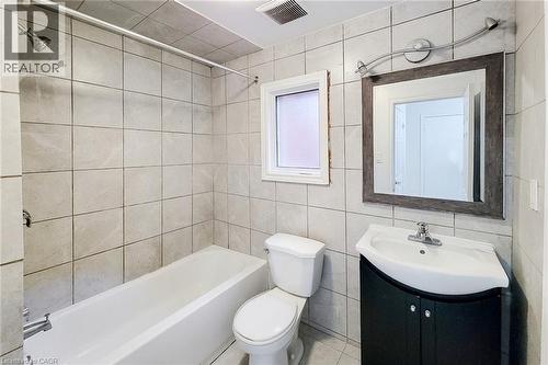 Bathroom featuring light-toned square wall and floor tiling, a white tub with shower fixture, a white toilet, a dark vanity with a white basin sink, and a framed mirror with track lighting - 463 Wellington Street N, Hamilton, ON - Indoor Photo Showing Bathroom