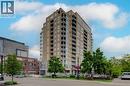Modern high-rise building featuring a light-colored brick exterior, expansive window walls, individual balconies, a ground-level commercial facade, and surrounding mature trees - 191 King Street S Unit# 607, Waterloo, ON 