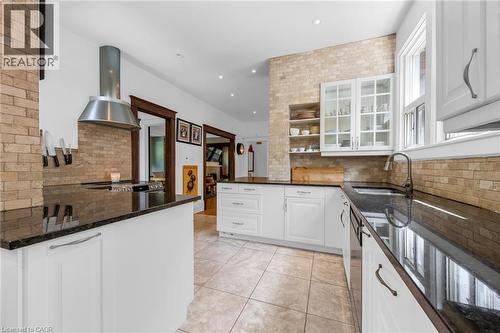Spacious kitchen featuring white cabinetry, dark stone countertops, a brick-style backsplash, a stainless steel range hood, and tile flooring - 110 Melrose Avenue S, Hamilton, ON 