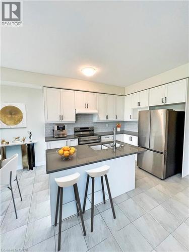 Modern kitchen featuring white cabinetry, stainless steel appliances, a dark countertop island with an undermount sink, a gray tiled backsplash, and light-toned tile flooring - 66 Aviron Crescent, Welland, ON 