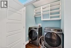 Dedicated laundry area featuring wood-finish flooring, two large capacity front-loading appliances, built-in open shelving, and a panel-style door - 
