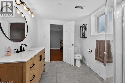 Bathroom featuring a wood-finish vanity with a white countertop and black hardware, a round black-framed mirror, a multi-bulb vanity light fixture, and a glass-enclosed shower - 25 Inverness Avenue, Hamilton, ON 