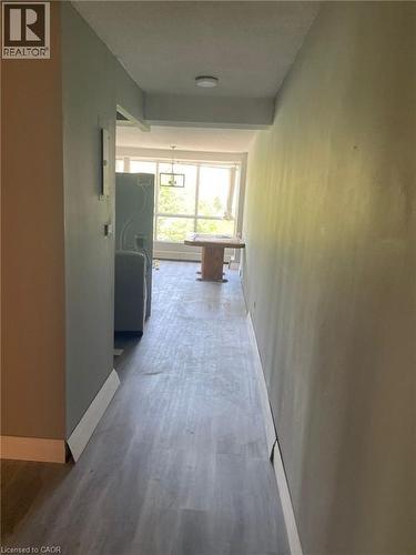 Hallway featuring wood-finish flooring, light-colored walls, and a view into a bright room with large windows and a contemporary ceiling light fixture - 150 Charlton Avenue E Unit# 708, Hamilton, ON 