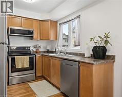 Kitchen featuring wood-finish flooring, light wood cabinetry, stainless steel appliances, a double basin sink with a window above, and stone-look countertops - 