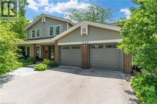Exterior featuring light gray siding with brick accents, an attached two-car garage with gray panel doors, a covered front porch with columns, a front entry door with sidelights, and a stone walkway - 1320 Monmouth Drive, Burlington, ON 