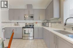 Kitchen featuring grey shaker-style cabinetry with brushed nickel hardware, light-toned countertops, a stainless steel electric range, an over-the-range microwave, and a double-basin sink with a goose - 